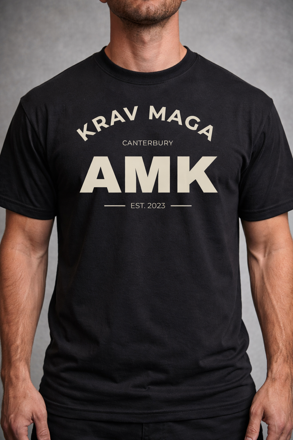 Person wearing a black  Krav Maga t-shirt with 'Krav Maga Canterbury AMK EST. 2023' text on a gray background